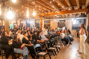 An audience seated in rows of chairs at an indoor event with exposed ceiling beams.
