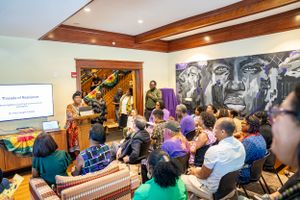 Crowded indoor presentation with audience members and speakers in front of dramatic wall artwork.