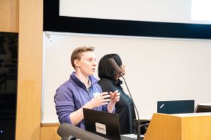 Two presenters speak into microphones during a professional conference presentation.
