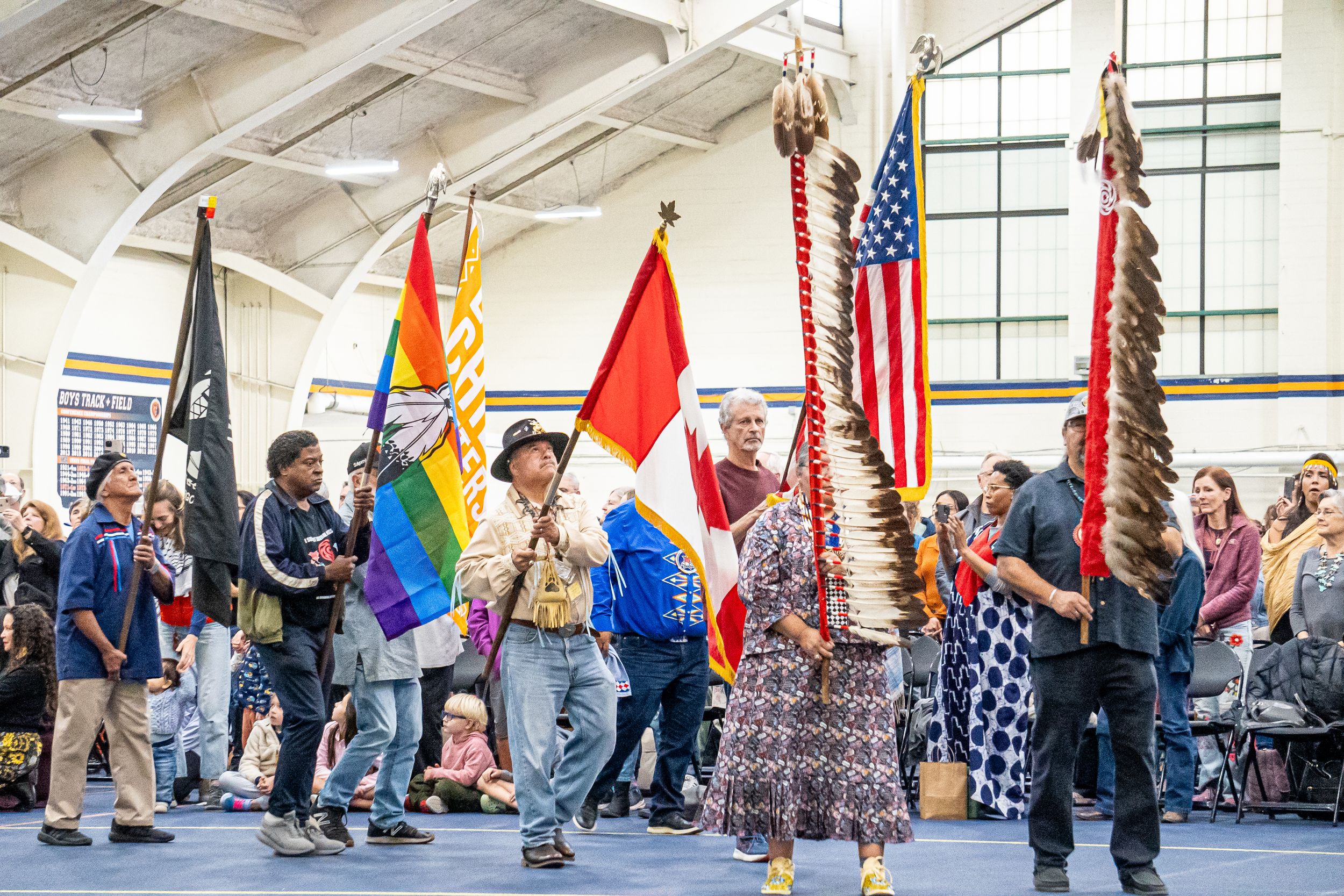 Flag bearers carry diverse national flags including American and Pride flags during an indoor ceremony with spectators.