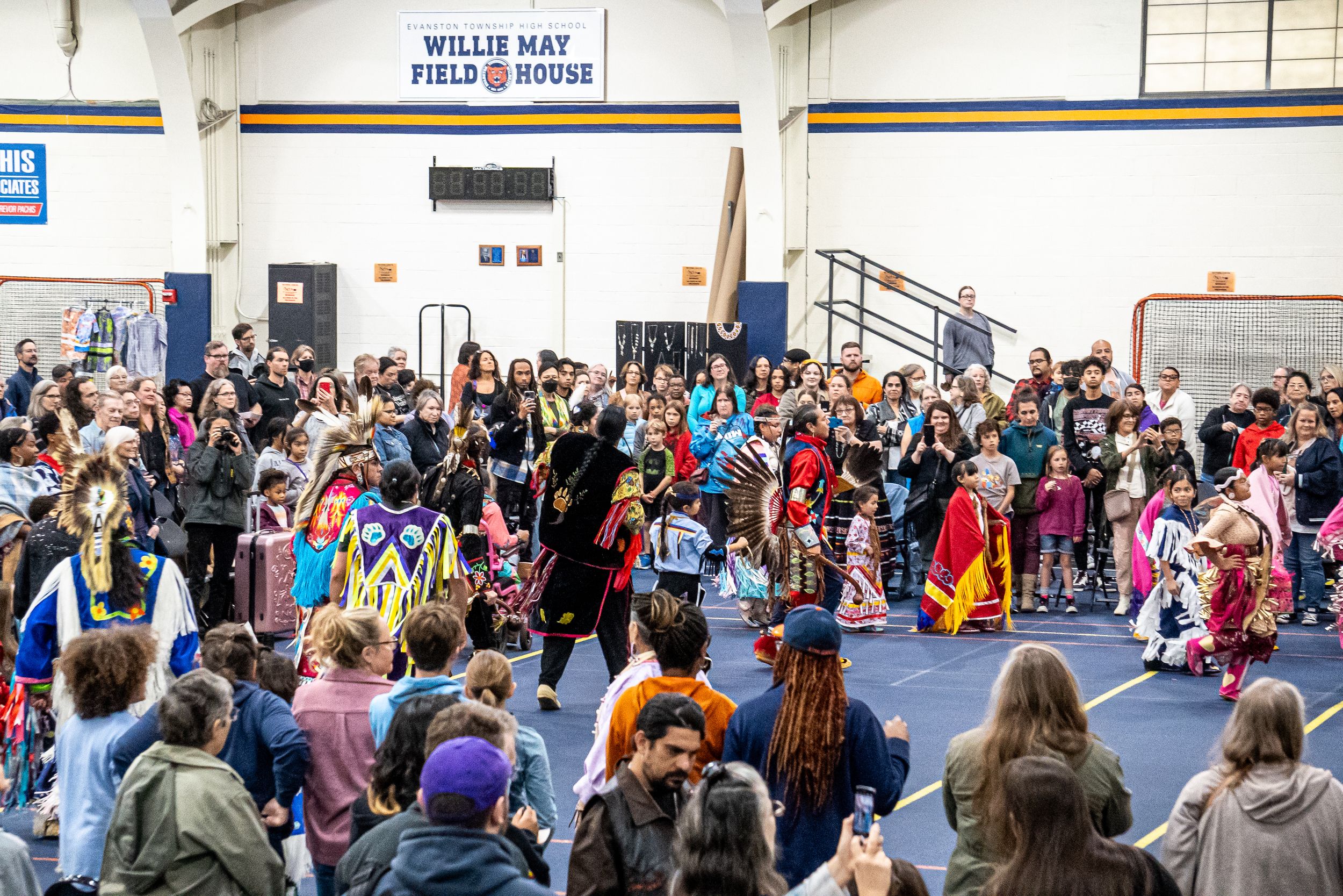 A large crowd watches a cultural performance in a school gymnasium with colorful costumes and raised hands.
