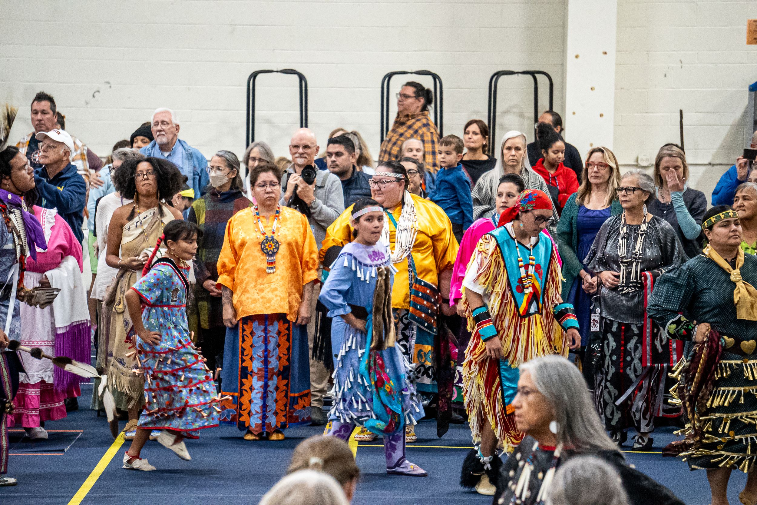 Colorful traditional Native American dancers gather at an indoor powwow celebration wearing vibrant regalia and ornate costumes.