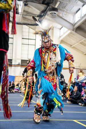 Traditional Native American dancer performs in vibrant blue and red regalia with feathered headdress at indoor powwow.