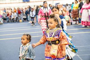 Pow wow dancers in traditional regalia perform on blue flooring at cultural celebration event.