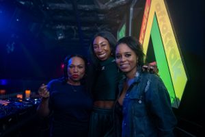 Three friends pose together in a dimly lit nightclub with colorful neon lighting in the background.