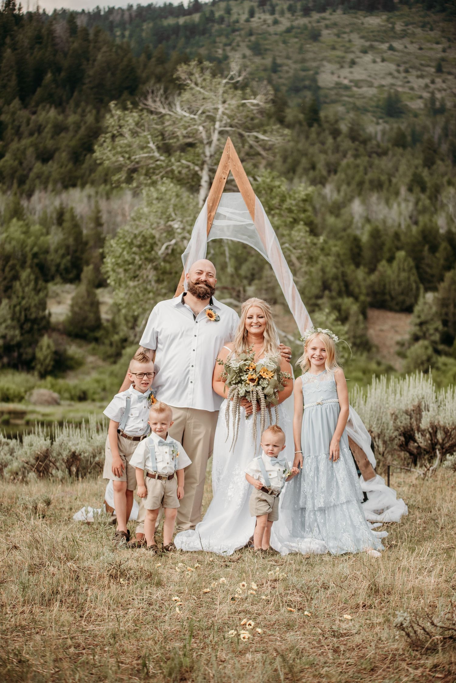 Wedding party poses in front of triangular arch in mountain setting with children.