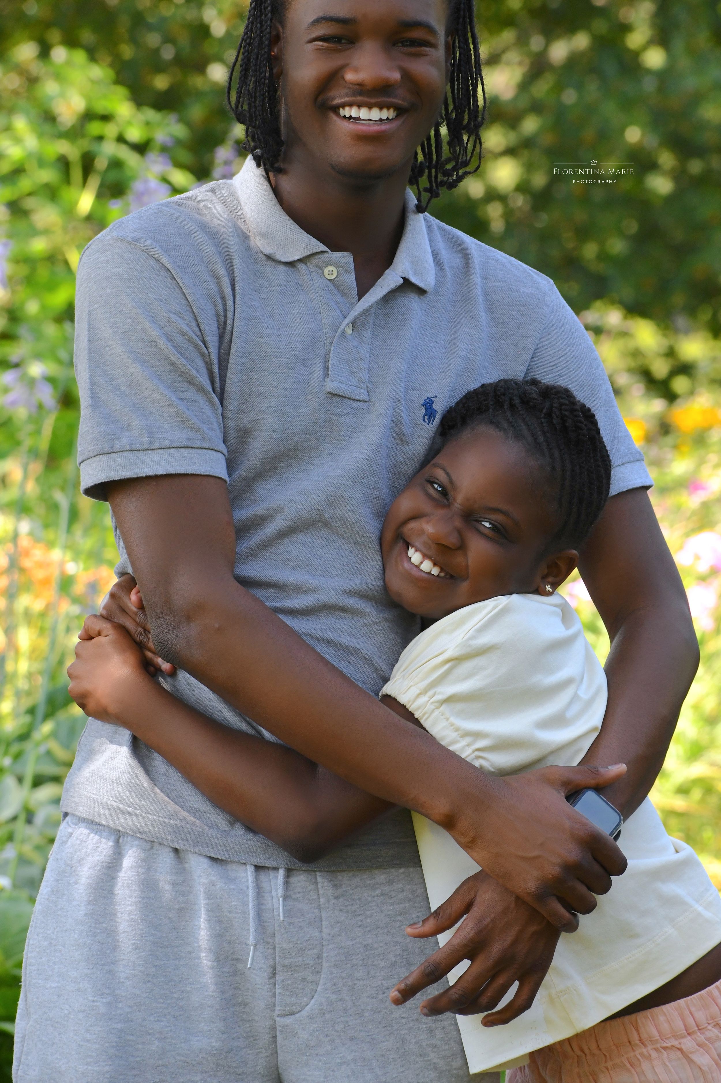 Two people embracing and smiling outdoors on a sunny day with flowers and greenery in the background.