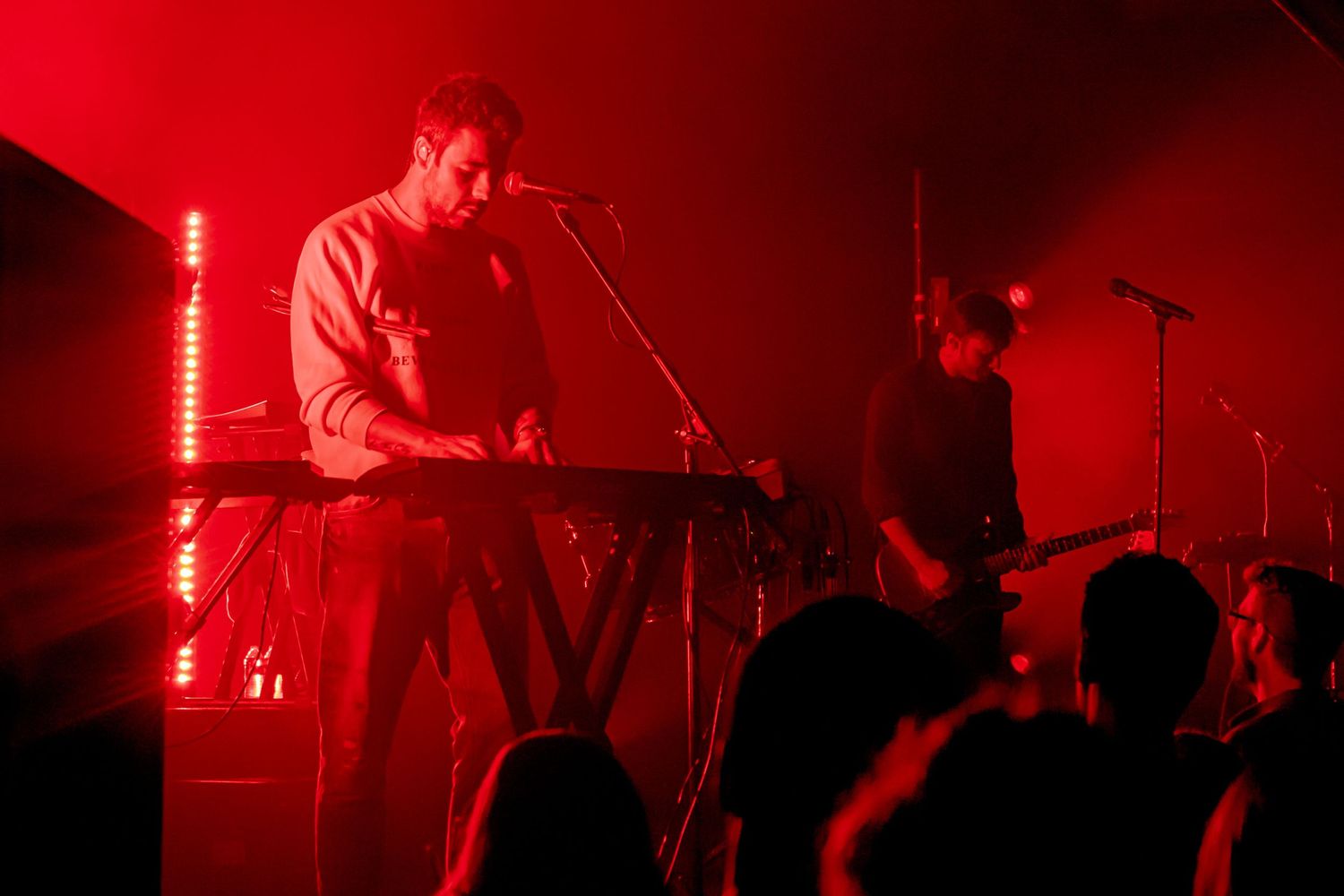 Musician performing on keyboard in dramatic red concert lighting while audience watches in silhouette.