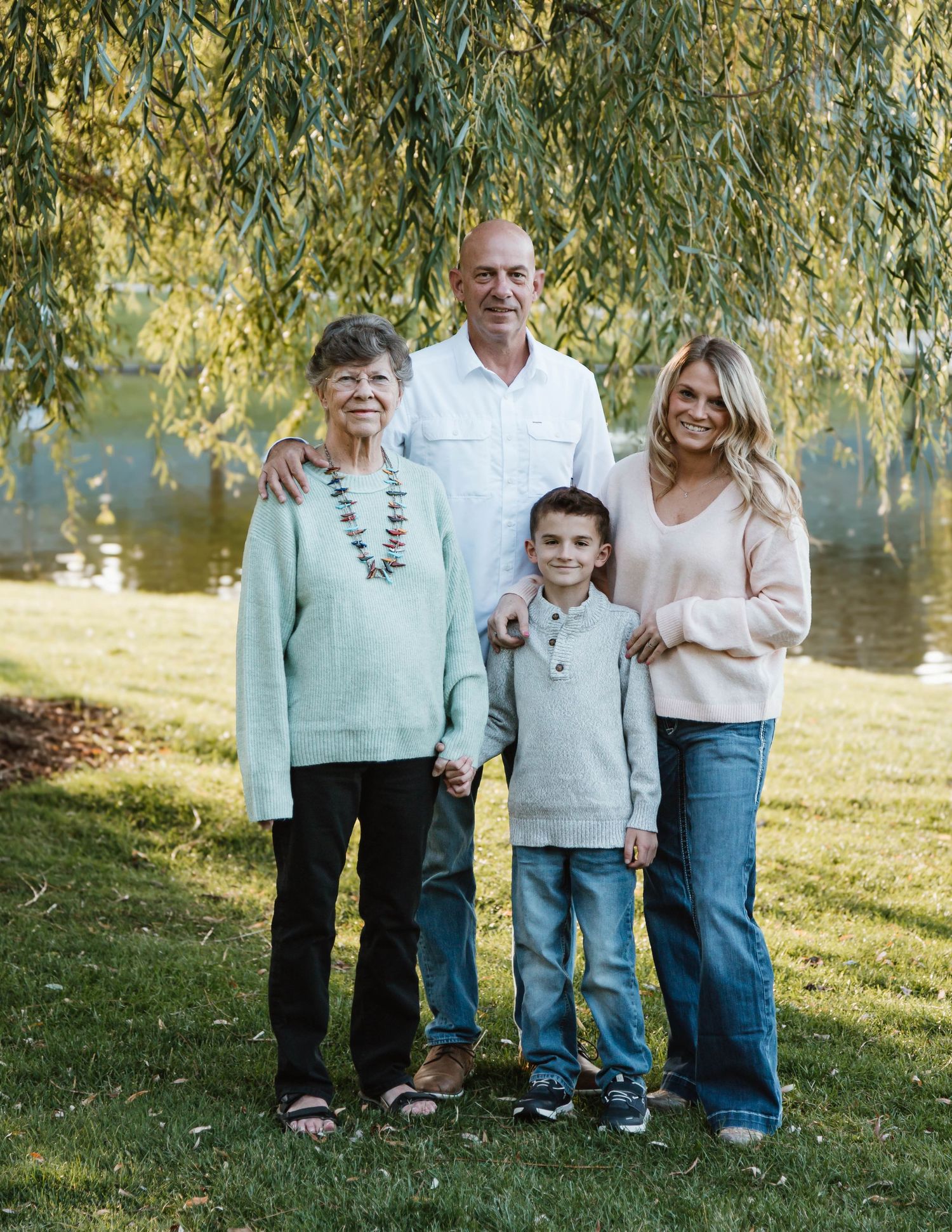 Four people standing together on grass under a willow tree near water on a sunny day.