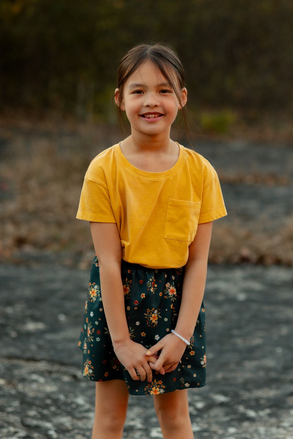 A portrait of a child in a yellow t-shirt and floral skirt standing on rocky ground.