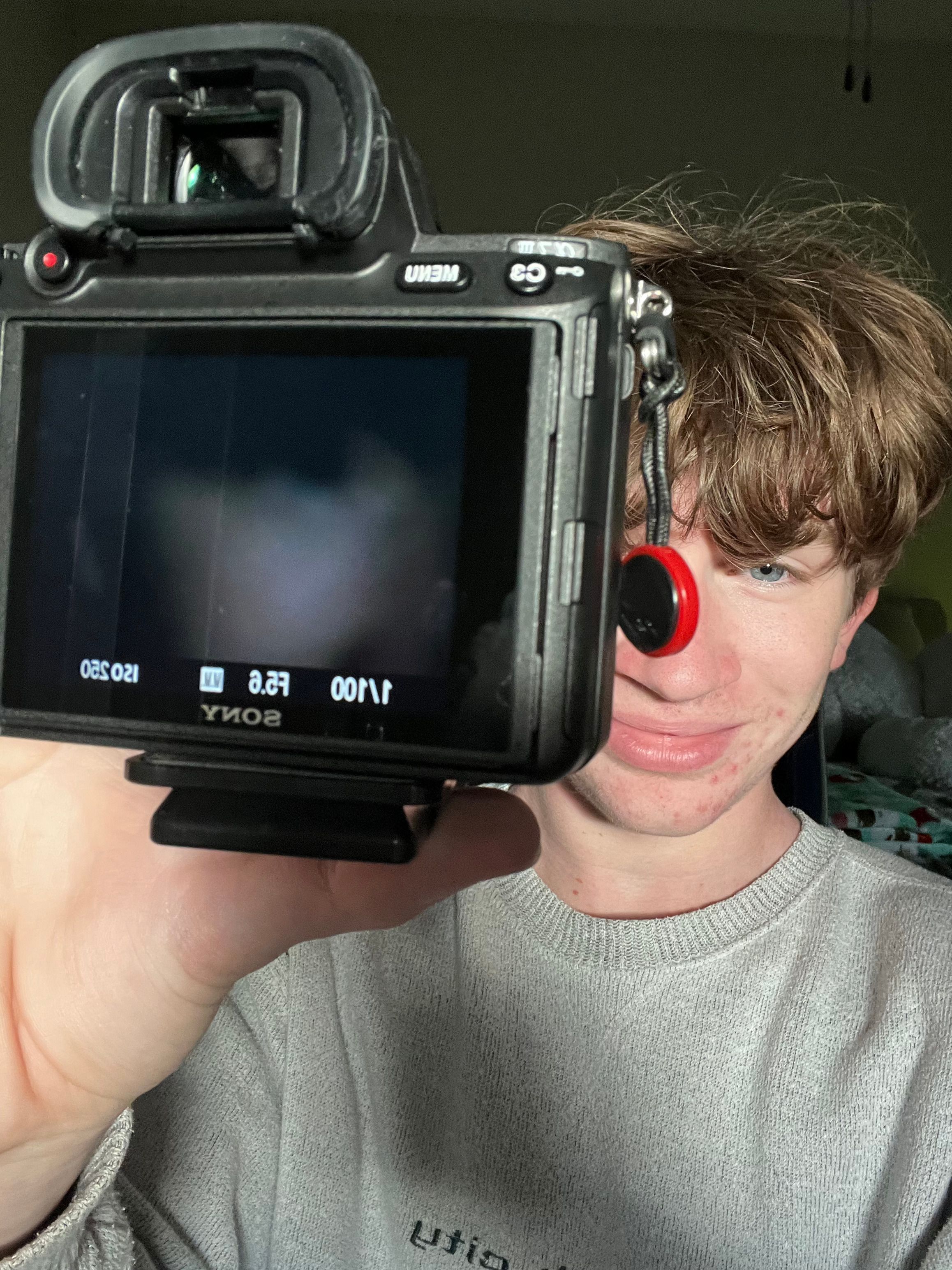 Young man holding a Sony mirrorless camera up to his face while taking a selfie indoors.