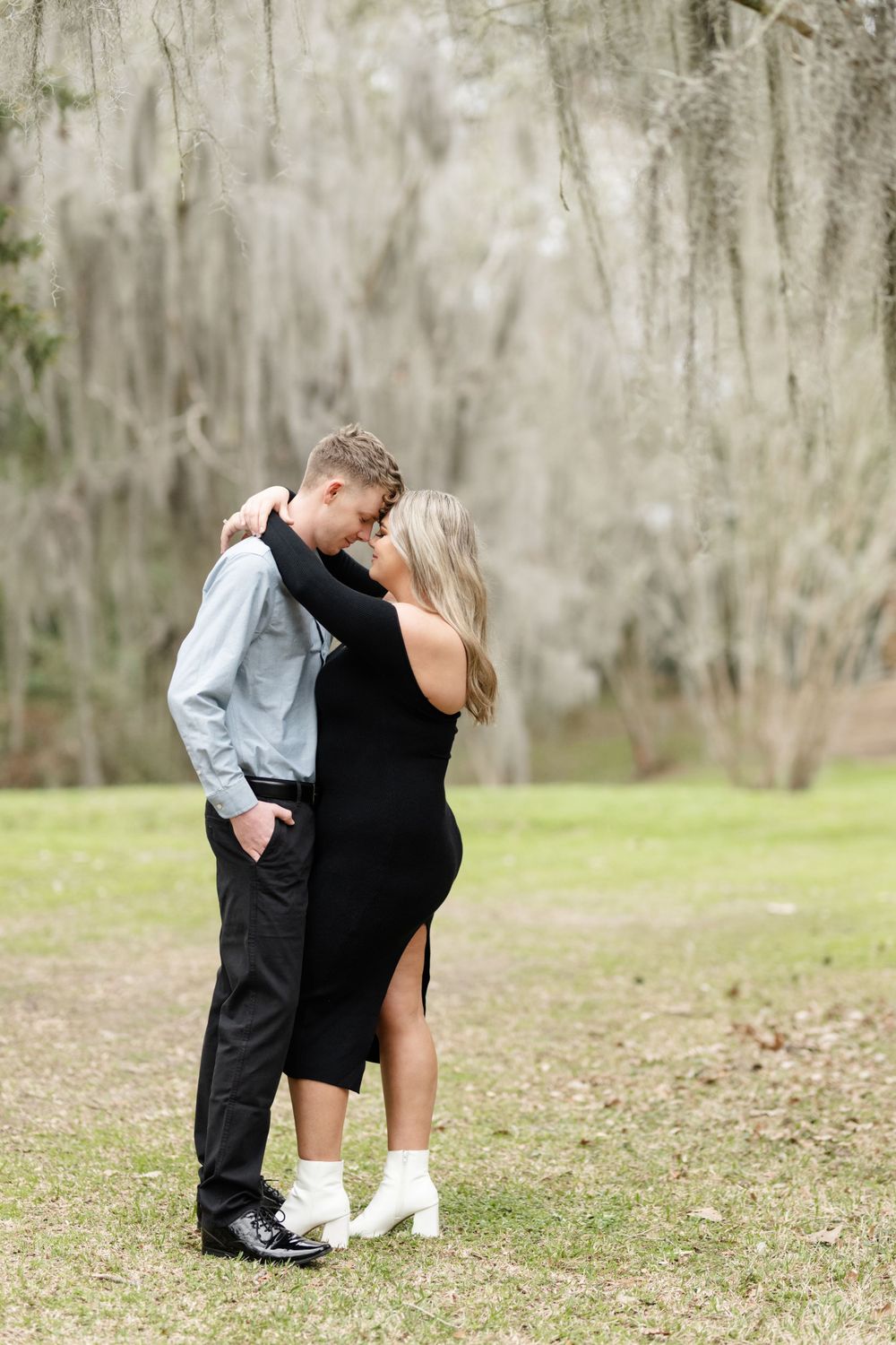 A couple shares an intimate moment outdoors under Spanish moss with her wearing a black dress and him in formal attire.