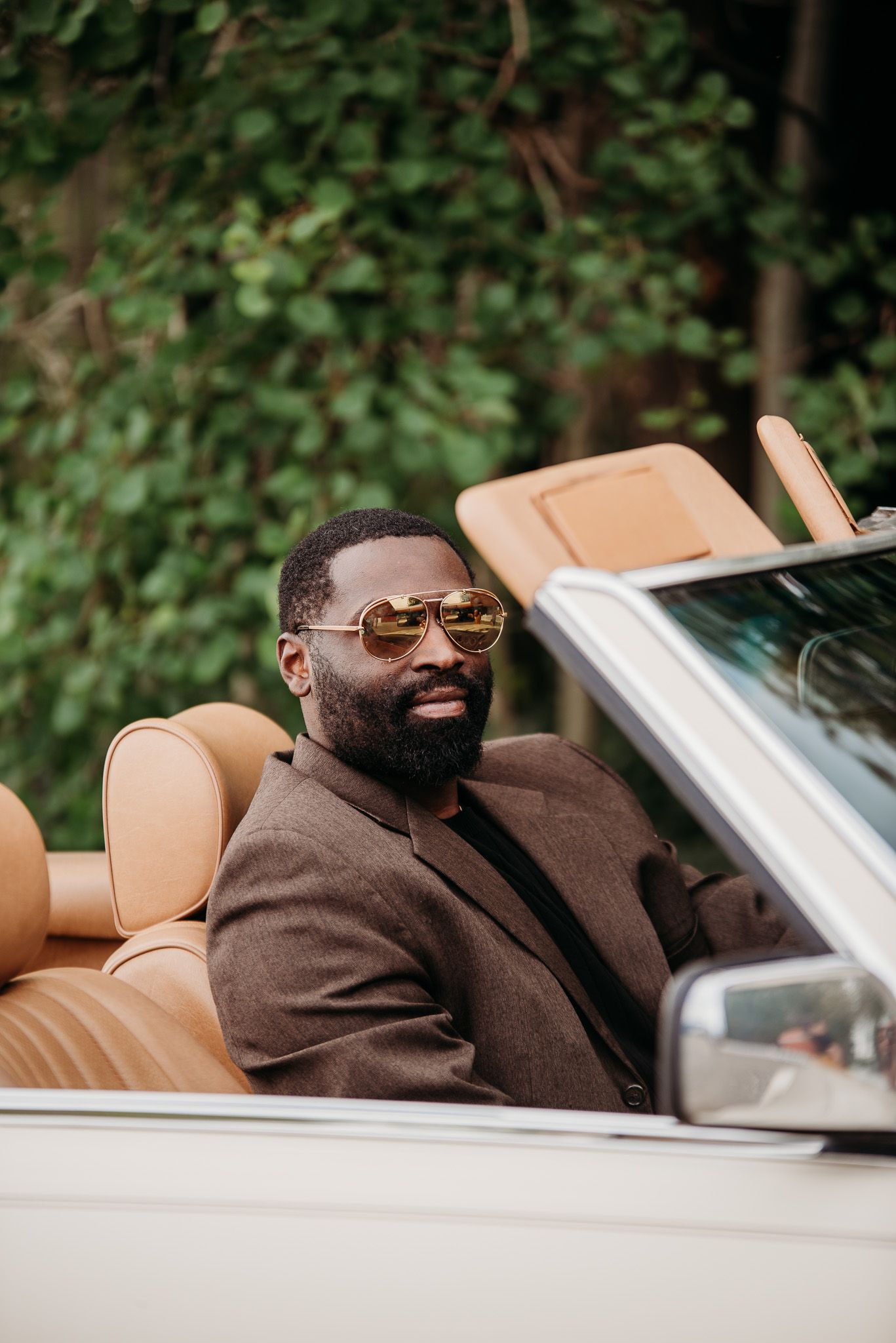 Stylishly dressed person in brown suit and sunglasses sitting in luxury convertible car with tan leather interior.