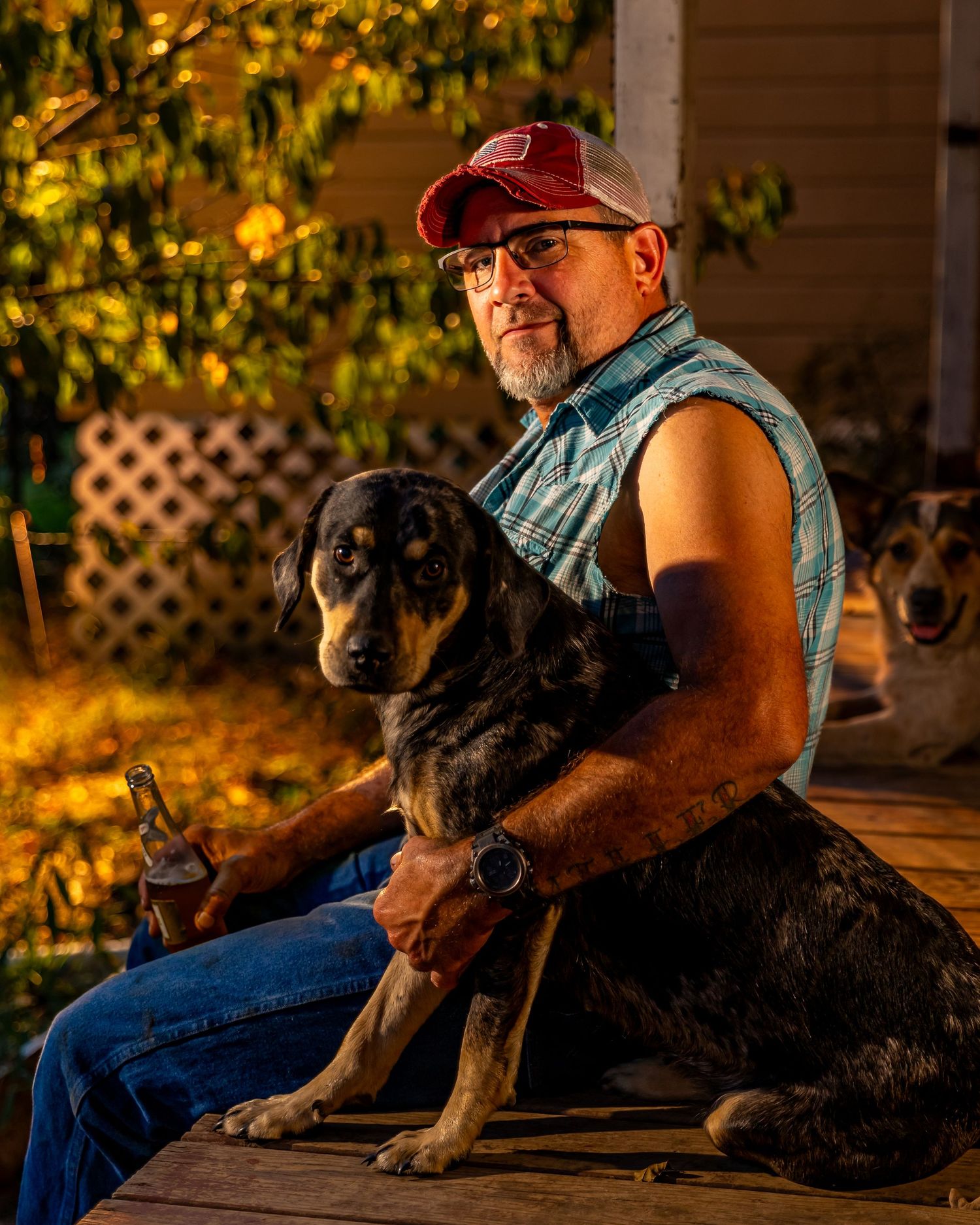 Seated person wearing red cap and denim vest holds a large black and brown hound dog on rustic porch at sunset.