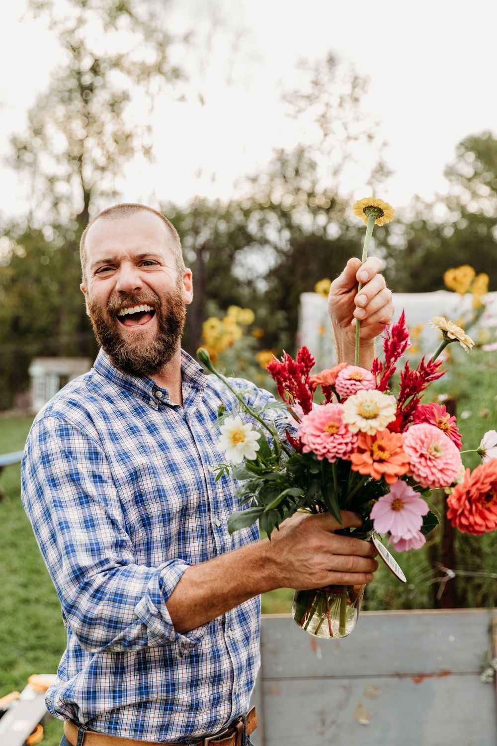 Man in blue plaid shirt arranging colorful summer flowers in rustic containers outdoors.