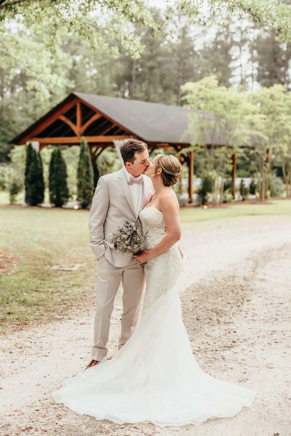 Romantic wedding portraits taken on a tree-lined path with rustic wooden structures in the background. North carolina wedding venue