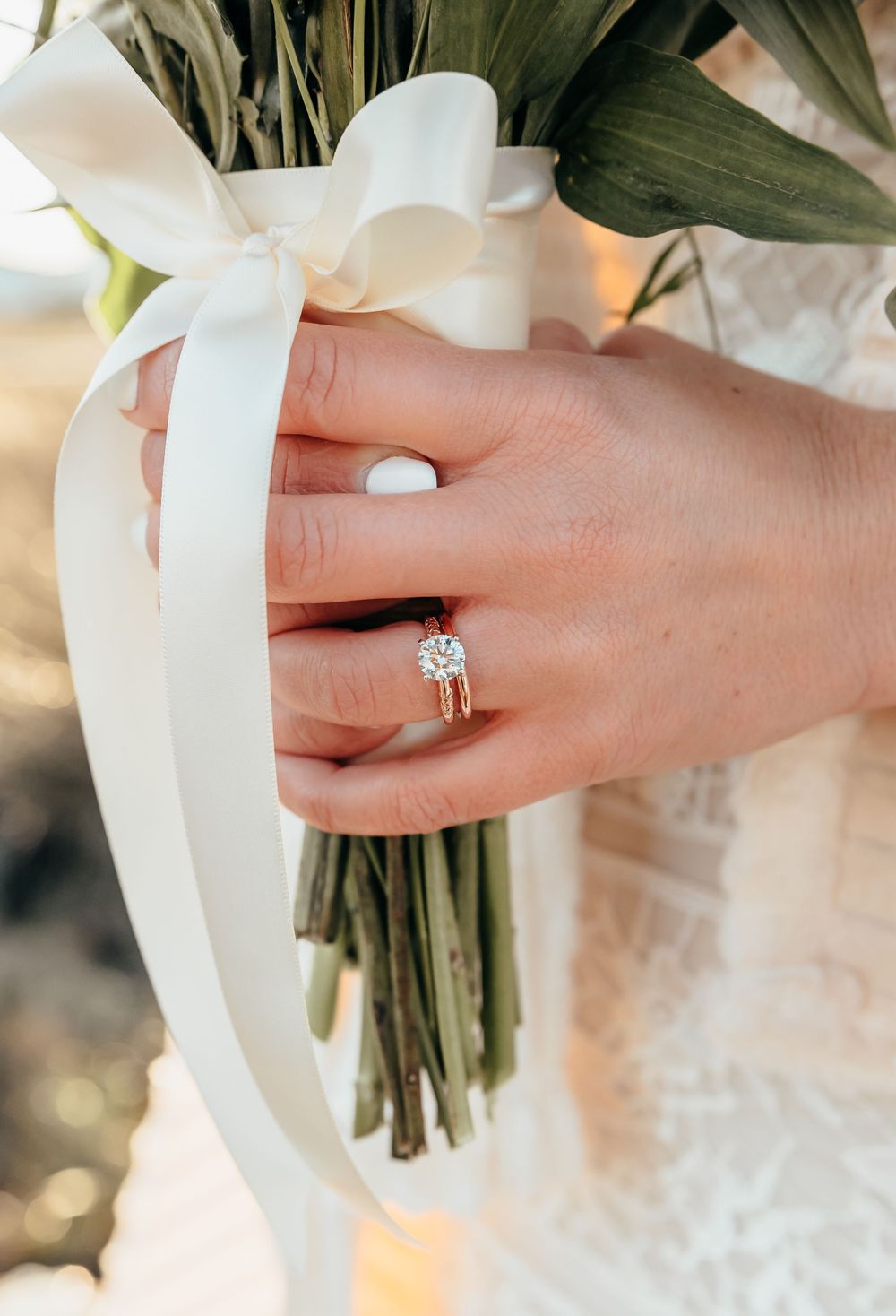 A close-up of delicate hands holding a bridal bouquet with white flowers wrapped in a satin ribbon.