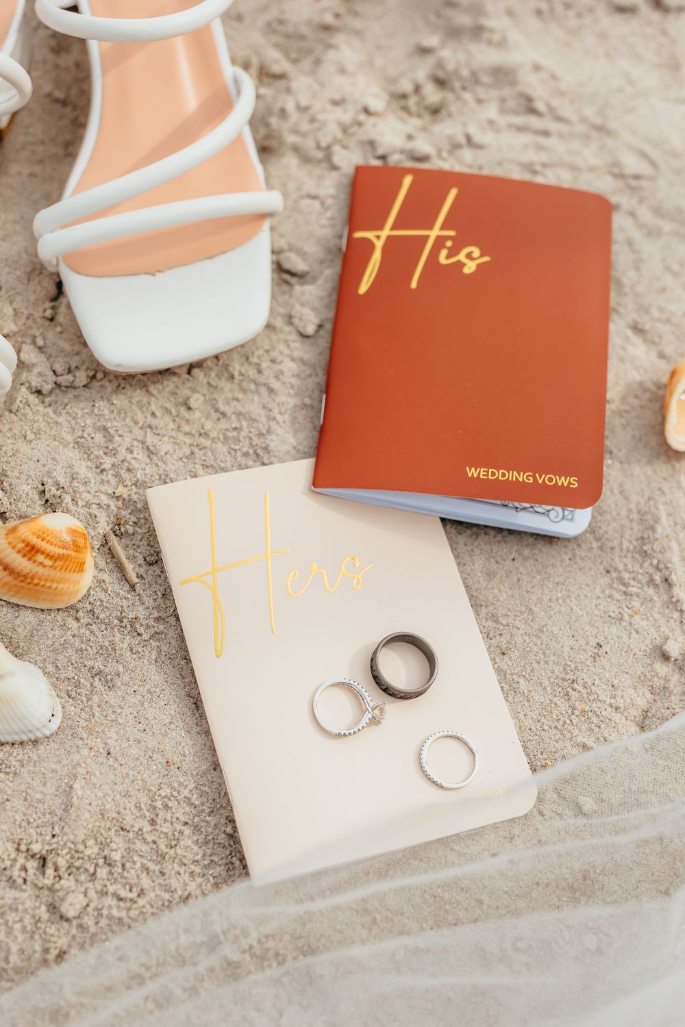 Wedding vow books in terracotta and cream colors displayed with white strappy sandals and wedding rings on sandy surface.