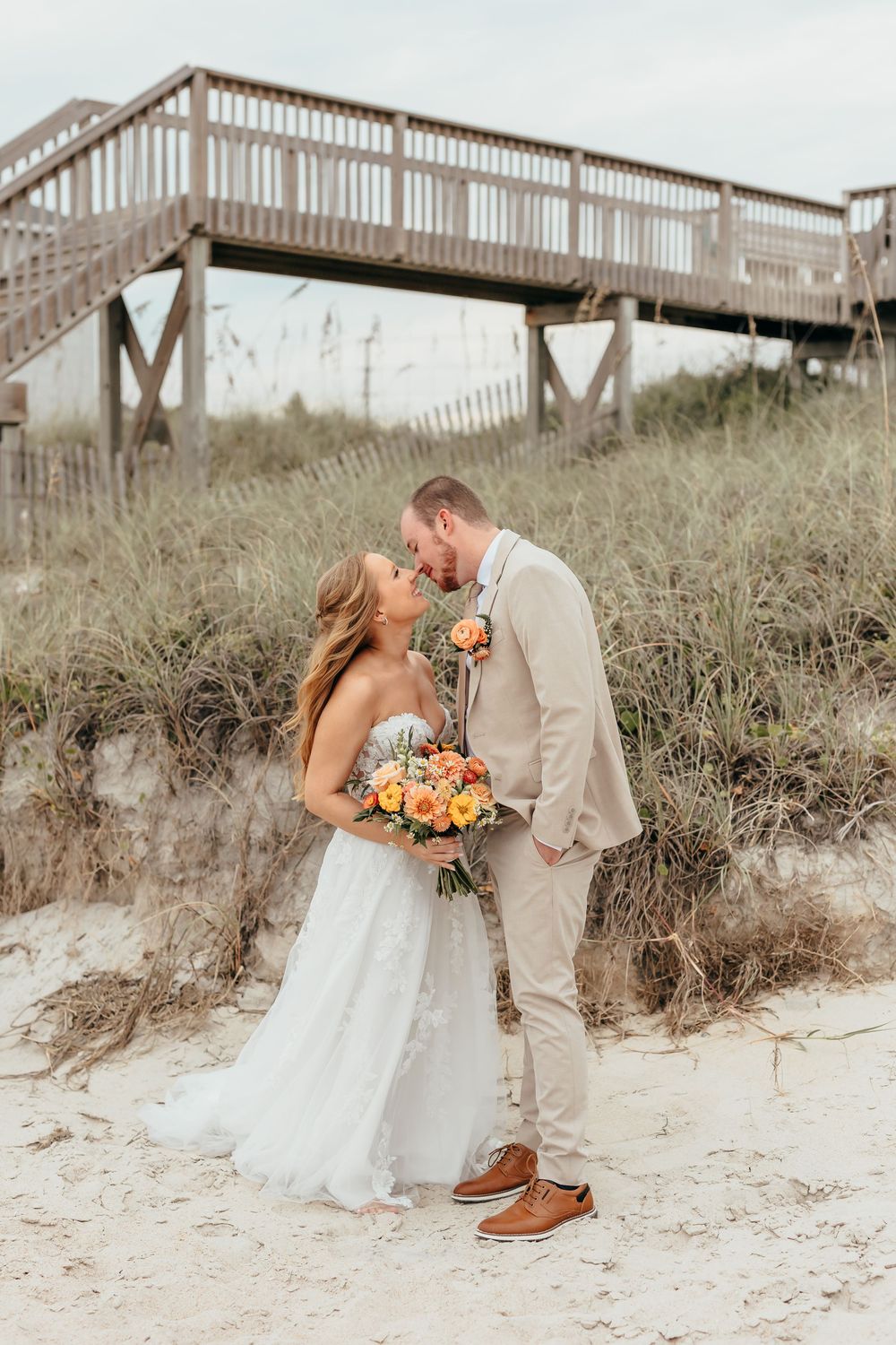 Romantic beach wedding photo shoot with couple sharing intimate moments on sandy dunes with wooden boardwalk backdrop.