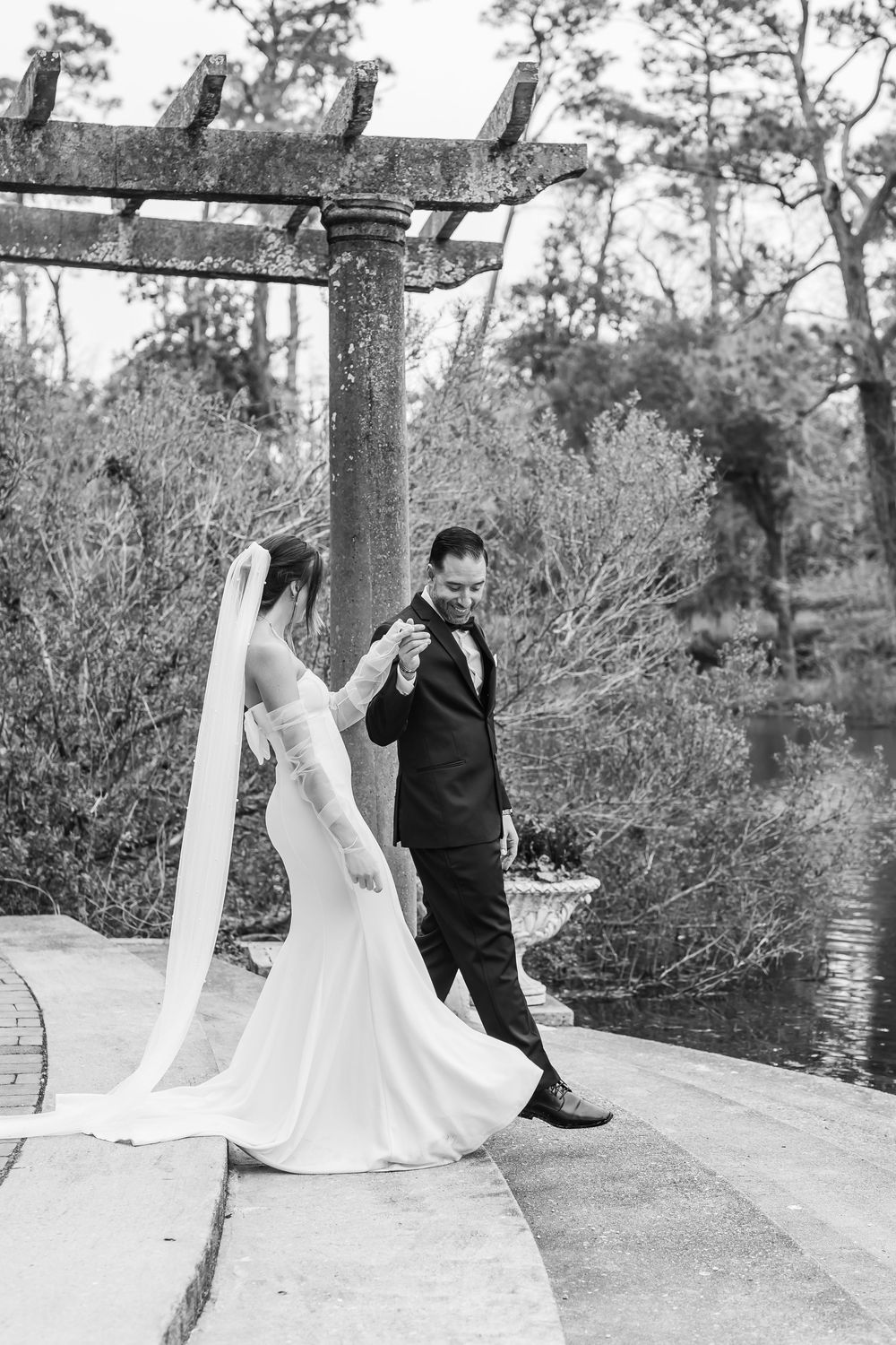 A black and white wedding portrait under a rustic pergola near water.