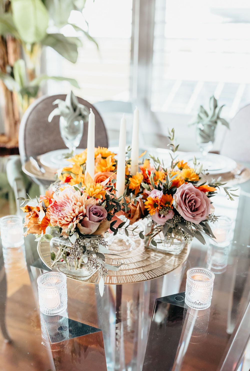 A romantic centerpiece with orange and mauve flowers surrounded by glowing candles on a glass table.