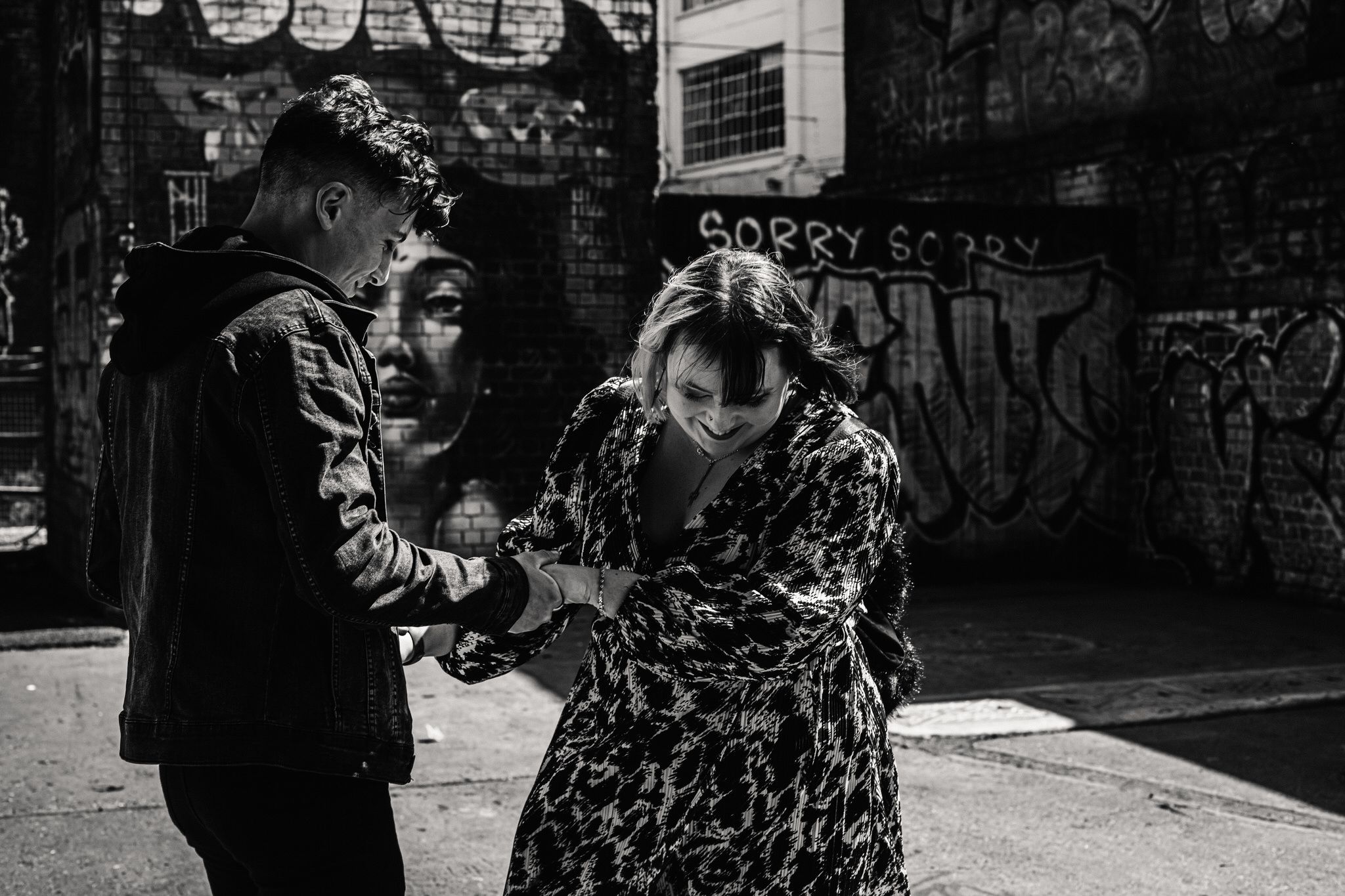 Two people share a candid moment on a city street in black and white with graffiti walls in the background.