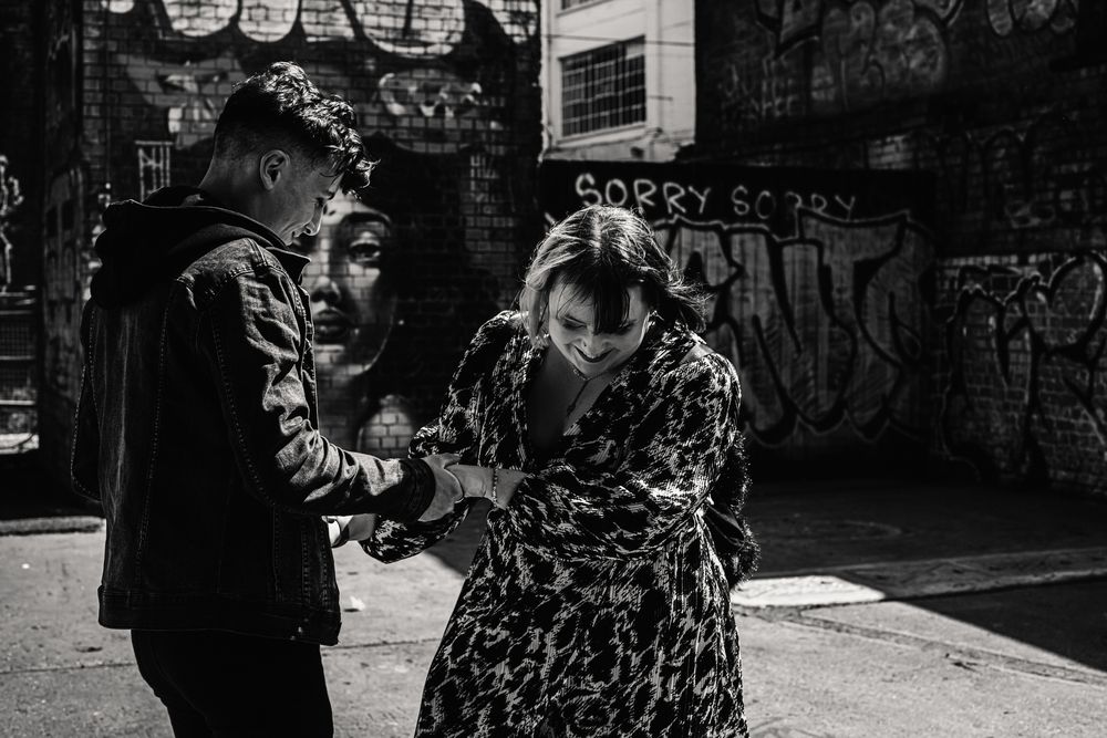 Two people share a candid moment on a city street in black and white with graffiti walls in the background.