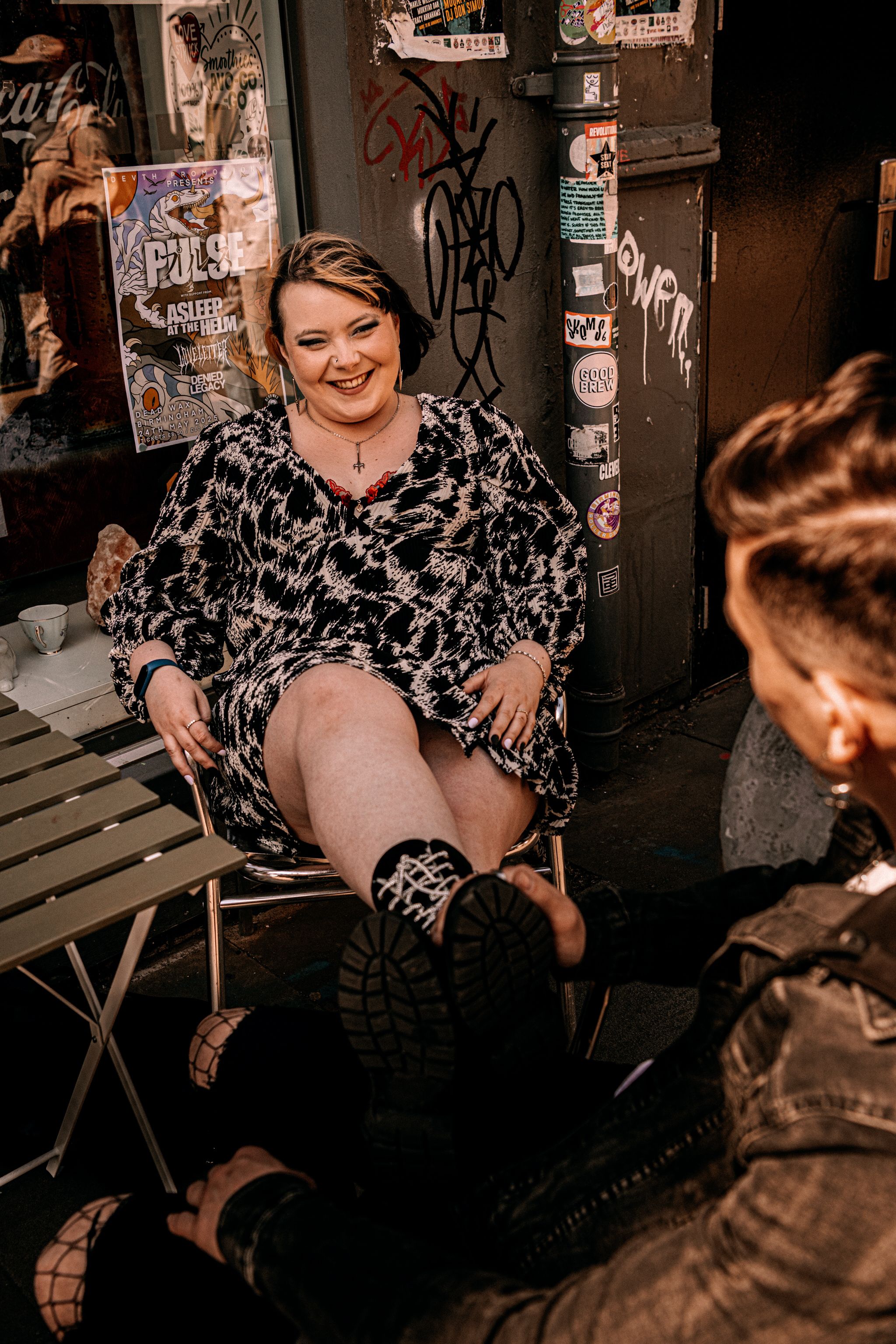 A person wearing a floral print dress sits on a folding chair against a graffiti-covered wall in a moody nighttime setting.