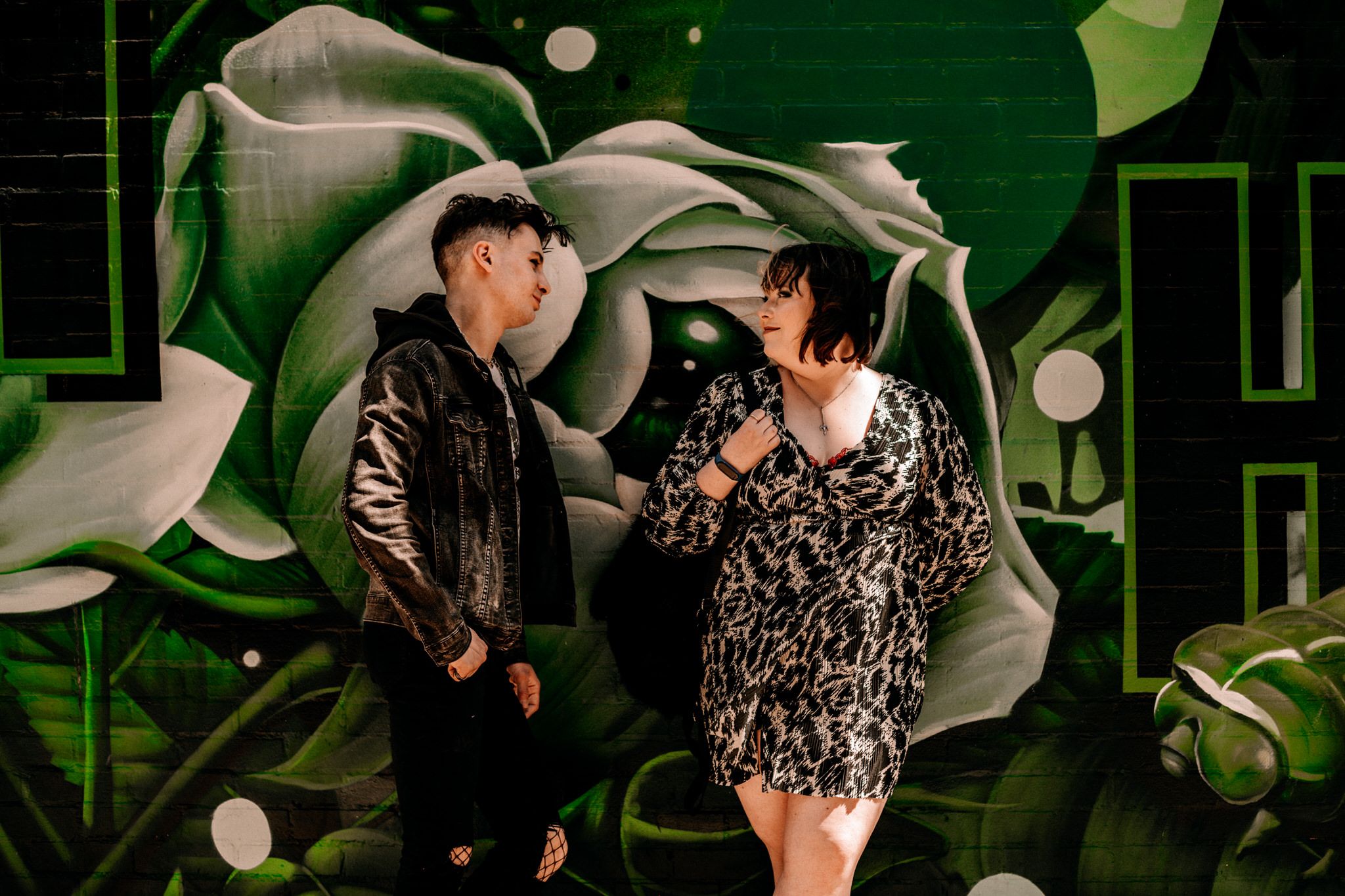 Couple poses playfully against vibrant green graffiti wall with floral designs at night.