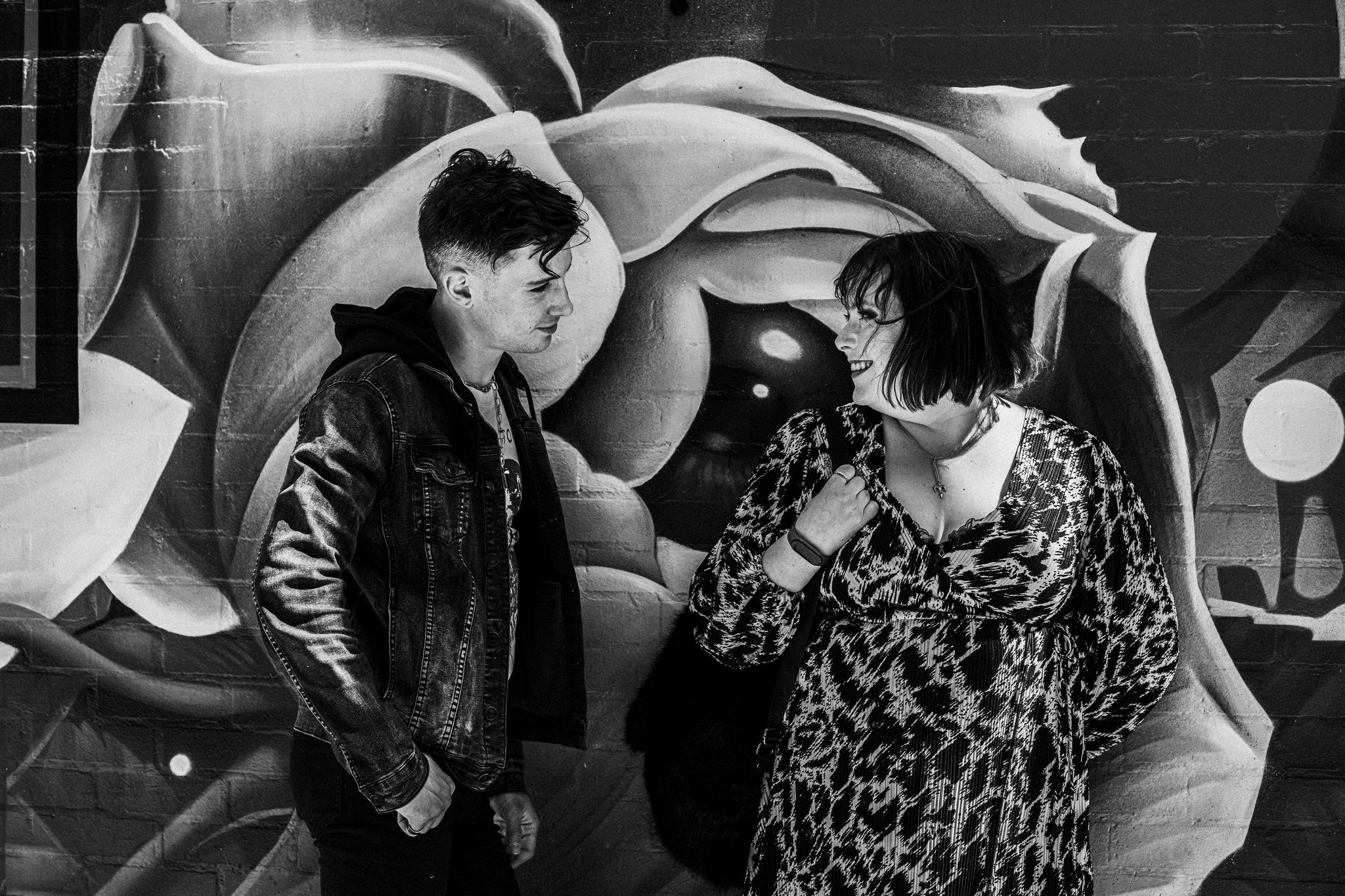 Two people conversing in front of a black and white graffiti mural wall with abstract designs.