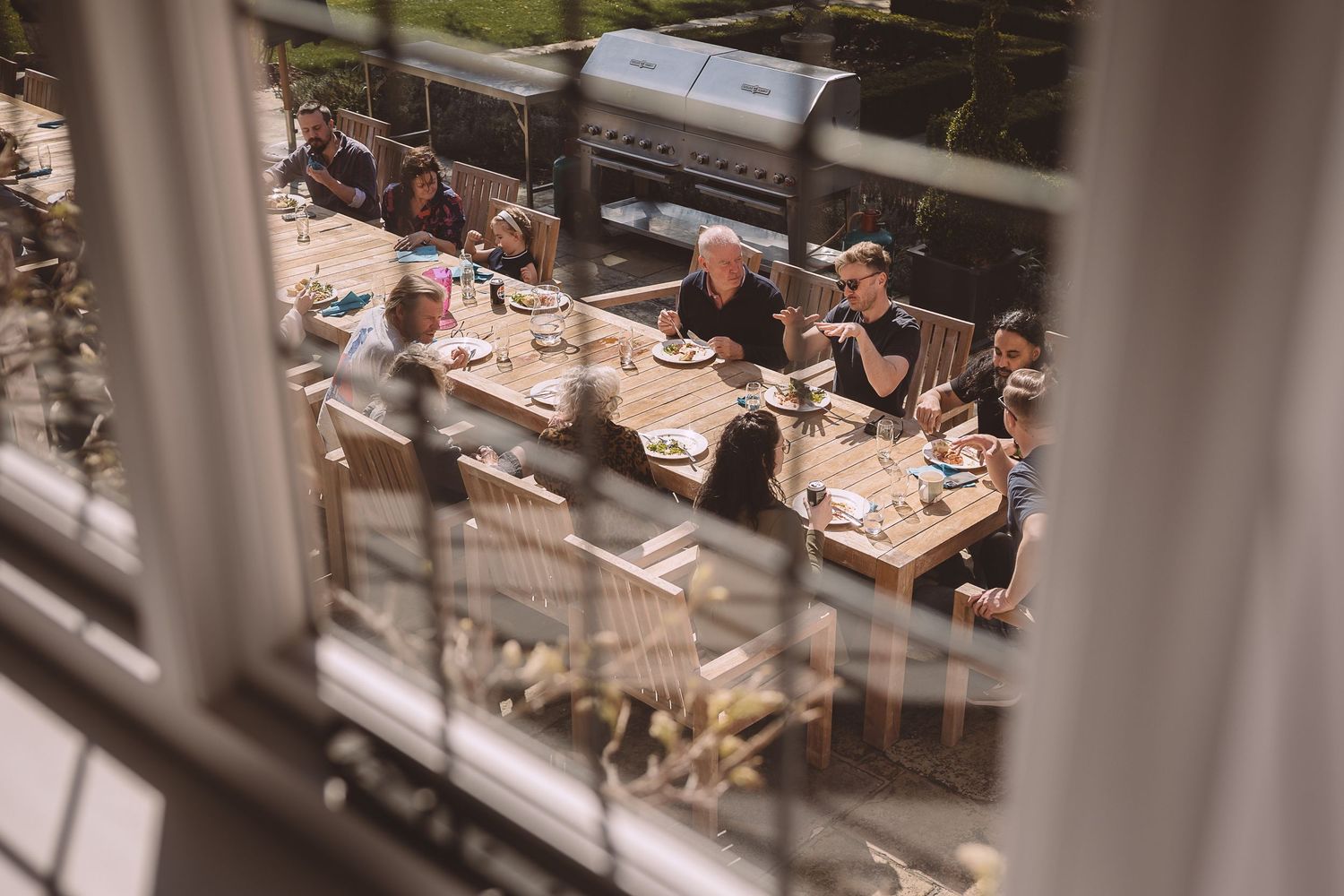 Guests enjoy outdoor dining at a restaurant patio viewed through window bars on a sunny day.