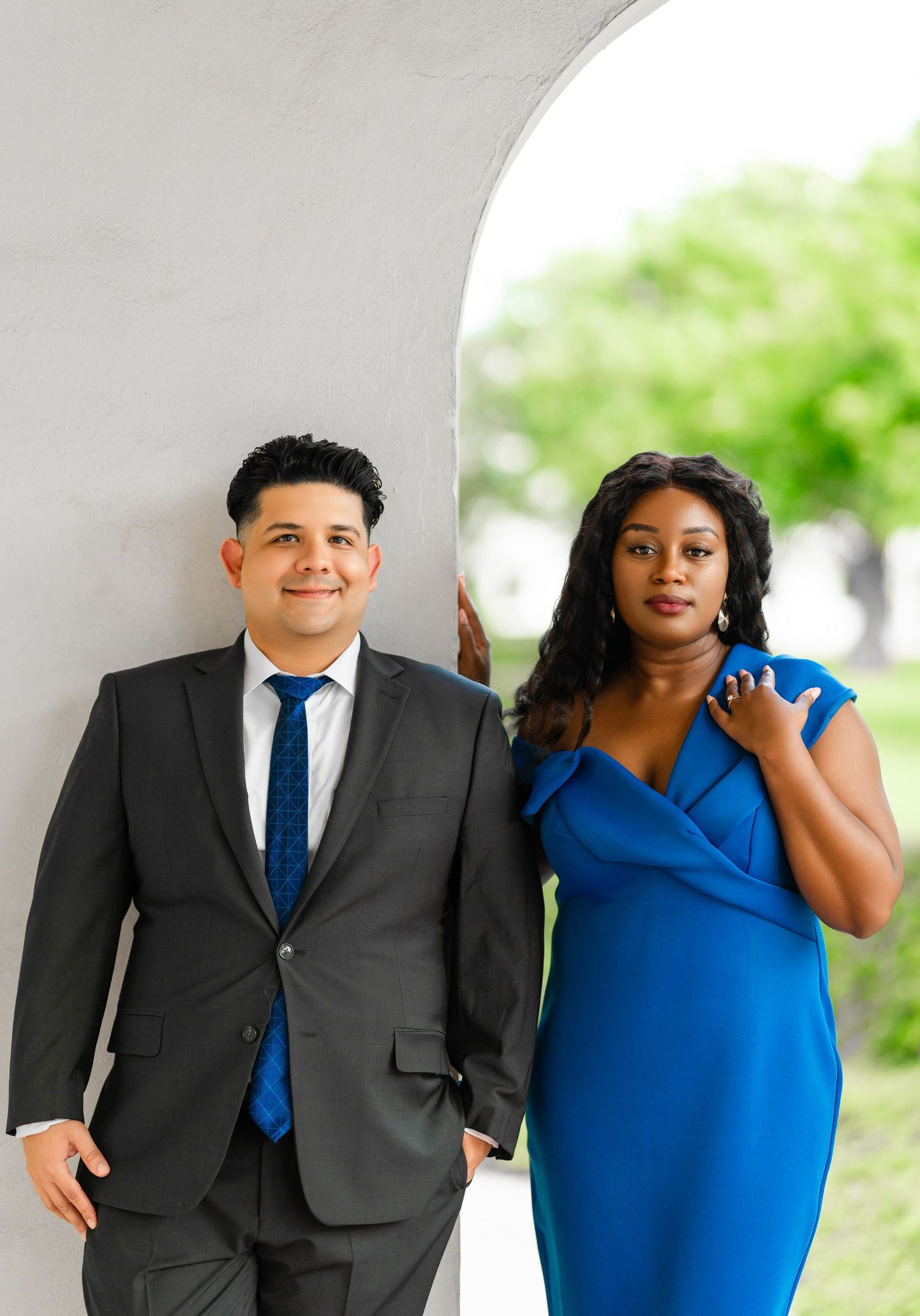 A couple in formal attire pose together under an archway, with the woman wearing blue and man in dark suit.