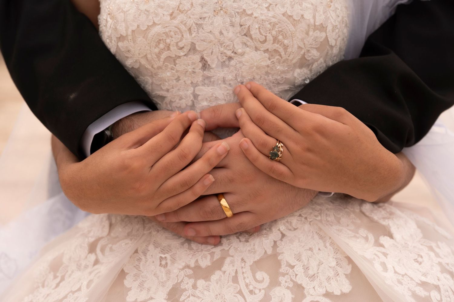 Close-up of two hands with wedding rings clasped together over a white lace dress.
