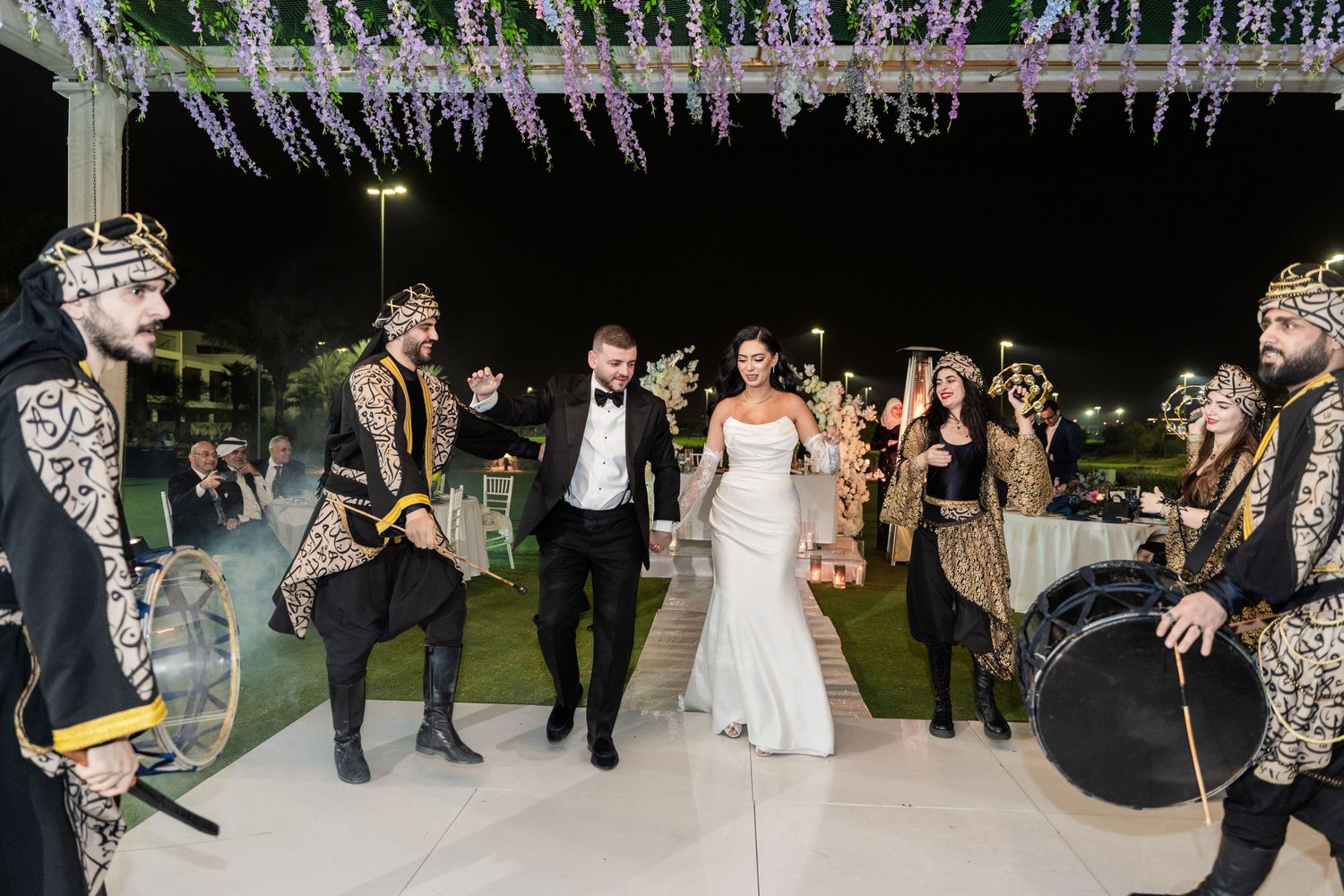 A celebratory wedding moment as newlyweds walk under twinkling lights while performers in elaborate costumes entertain guests.
