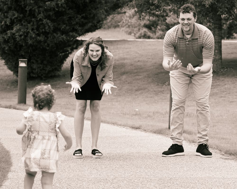 Black and white photo of adults outdoors playing with a small toddler in a dress on a park path.