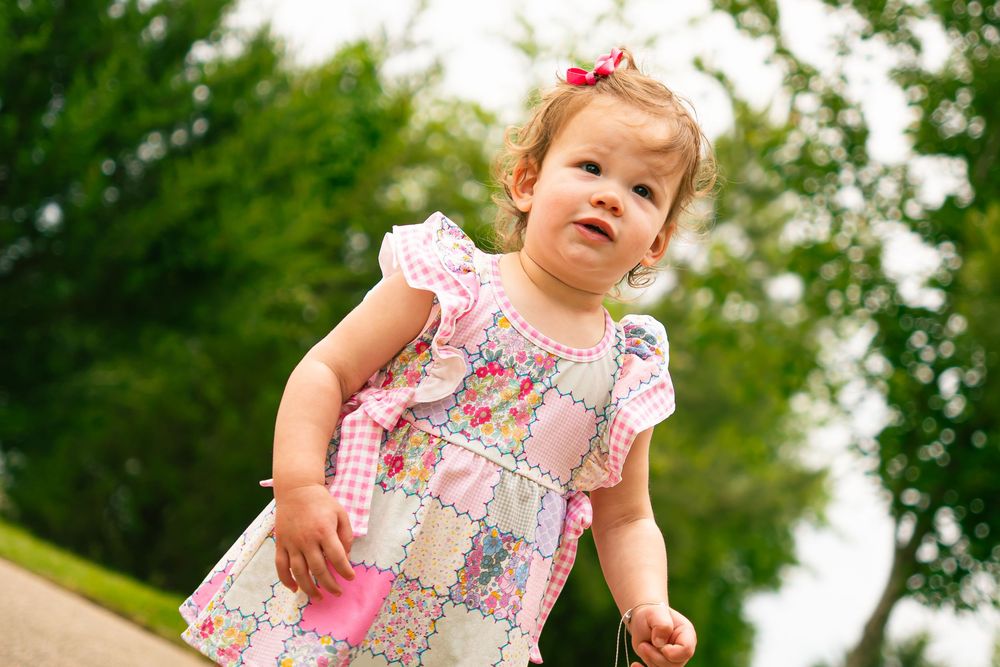 Small child wearing a floral dress with pink bow standing outdoors against green foliage background.