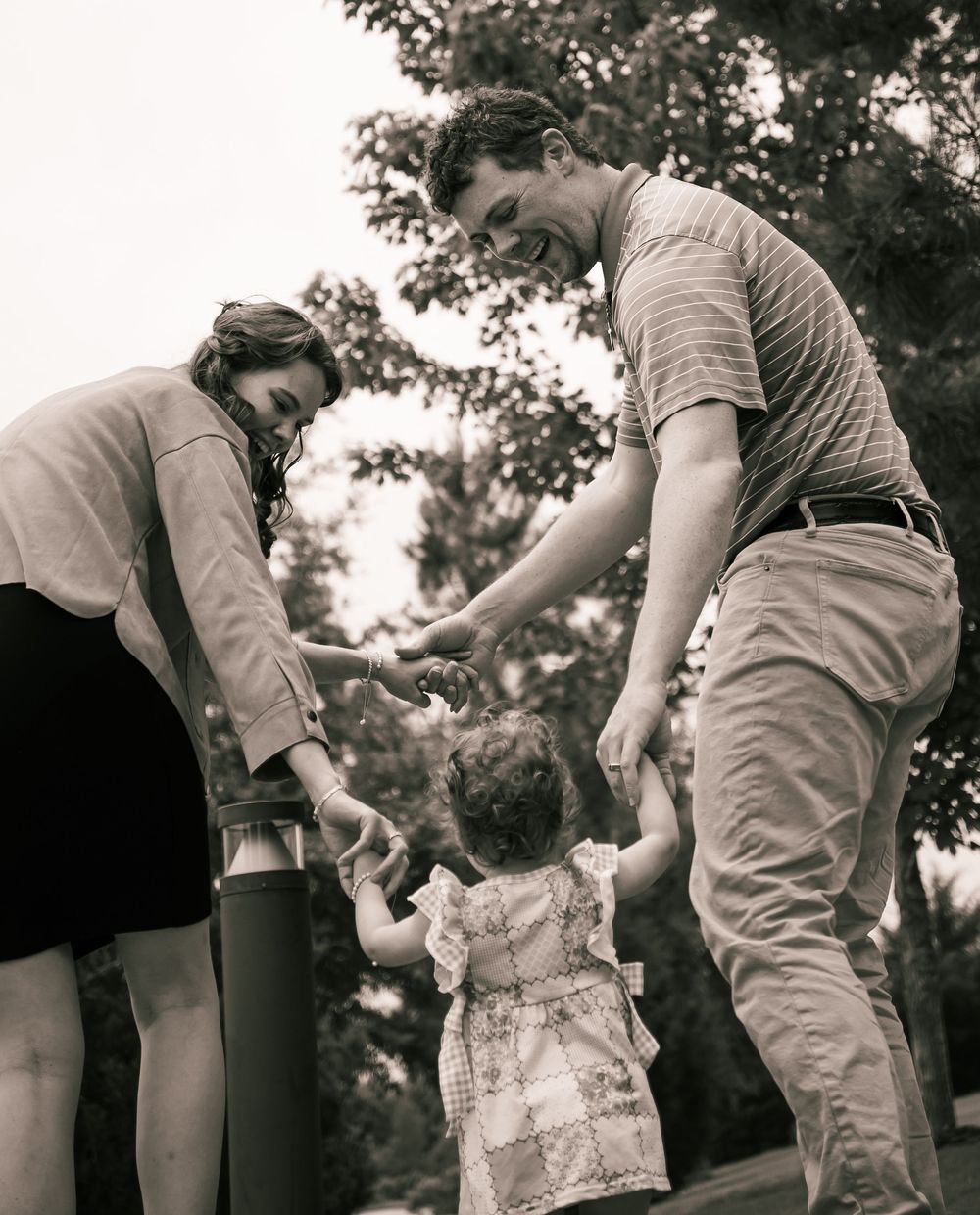 A vintage black and white photograph of a family holding hands with a toddler in a dress outdoors.