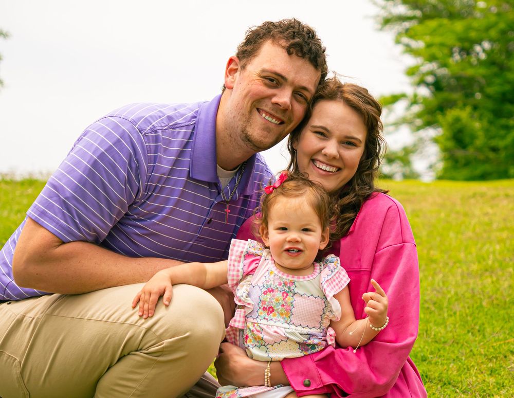 A family sits together on a grassy hill on a bright summer day, smiling warmly at the camera.