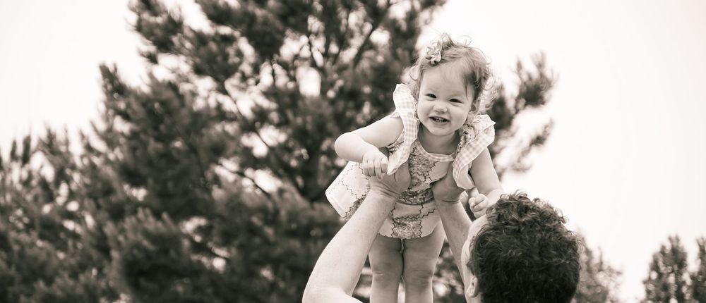 A playful black and white photograph showing someone lifting up a smiling toddler against a backdrop of trees.