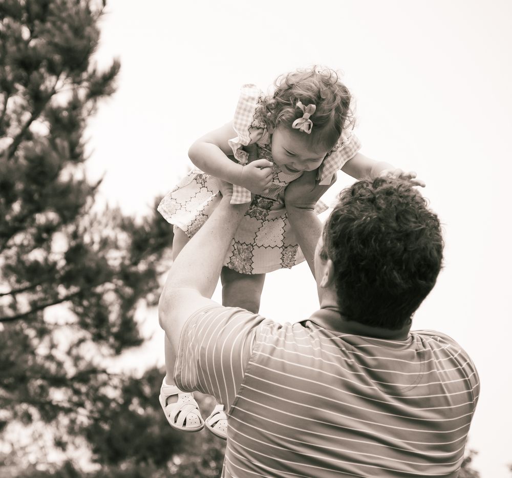 A parent lifts a small child wearing a checkered vest up in the air during a tender black and white moment outdoors.