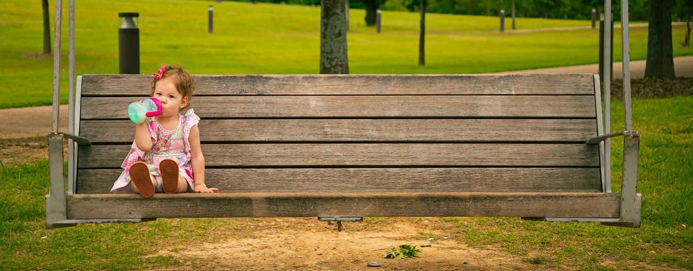Small child sitting alone on a large wooden swing in a scenic park setting with green grass.