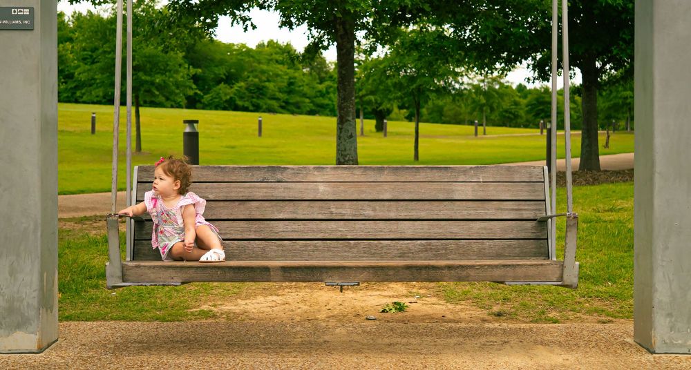 Two small figures sit together on a wooden park swing suspended between concrete pillars in a green summer setting.