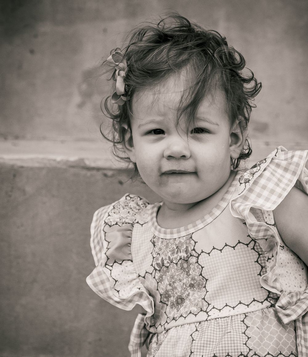 A black and white portrait of a curly-haired toddler in a patterned dress smiling at the camera.