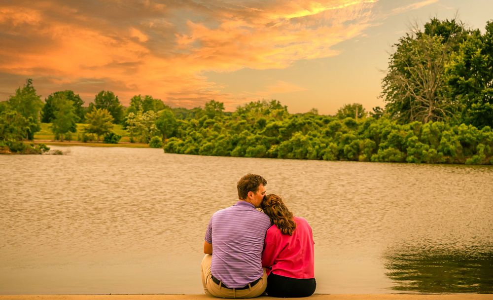 Two people sit close together on a riverbank at sunset while orange and pink clouds reflect on the calm water.
