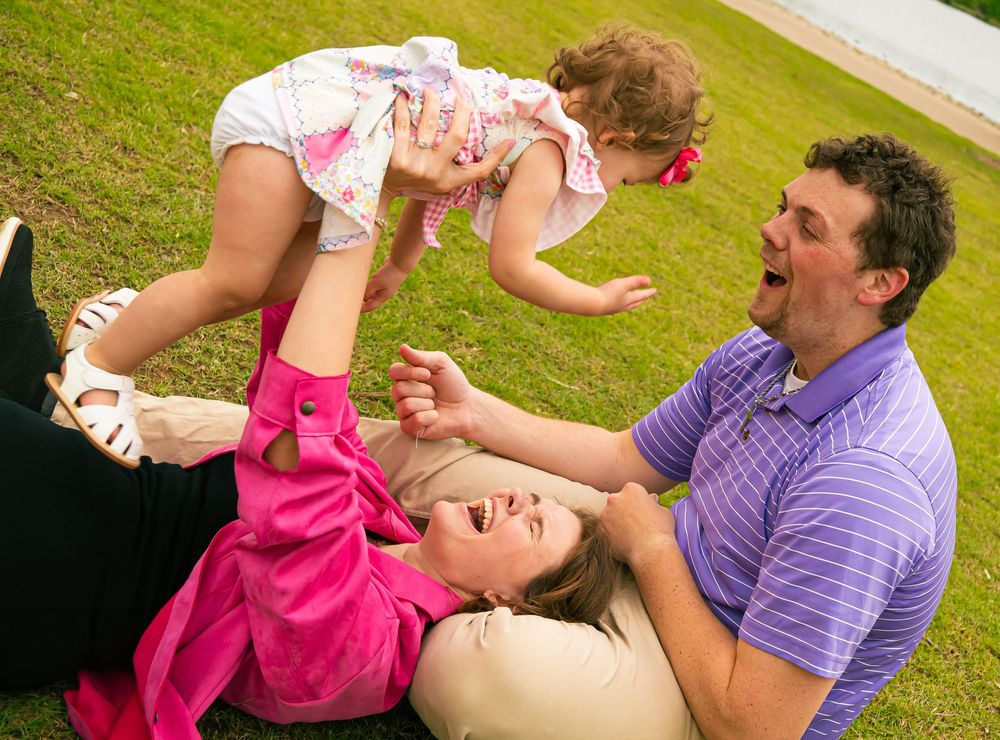Family lounging on grass together playfully lifting little one into the air during outdoor playtime.