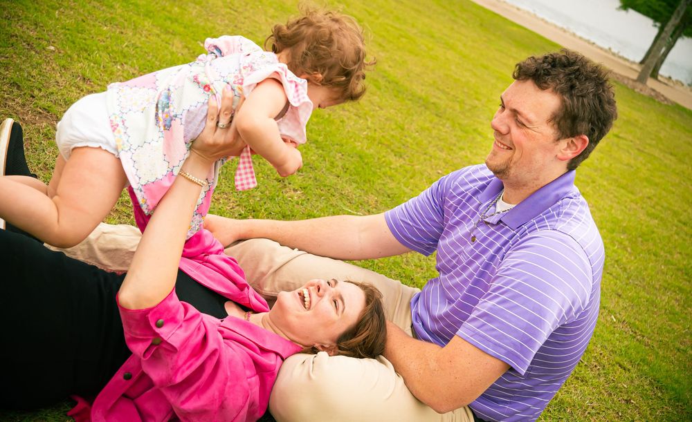 A group of three people play and laugh together on a grassy lawn on a sunny day.