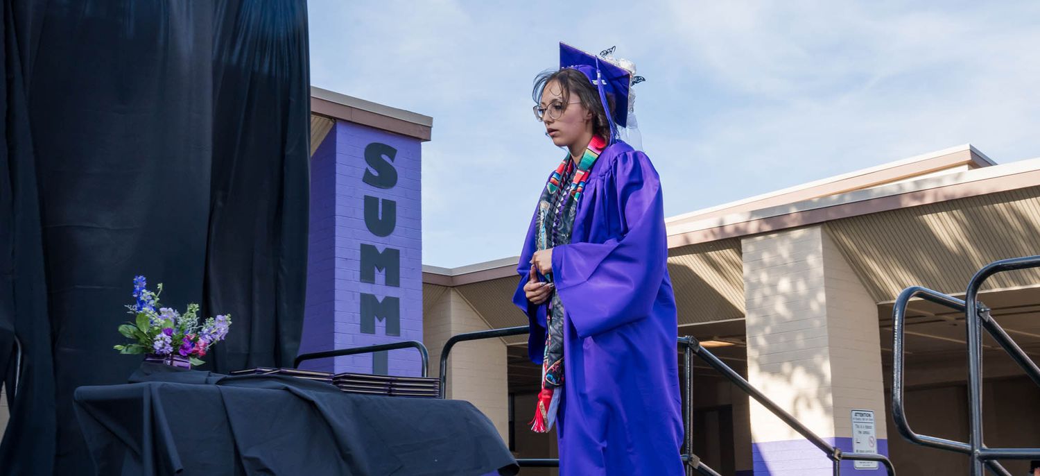 Graduate in blue cap and gown stands on outdoor stage during commencement ceremony under sunny sky.