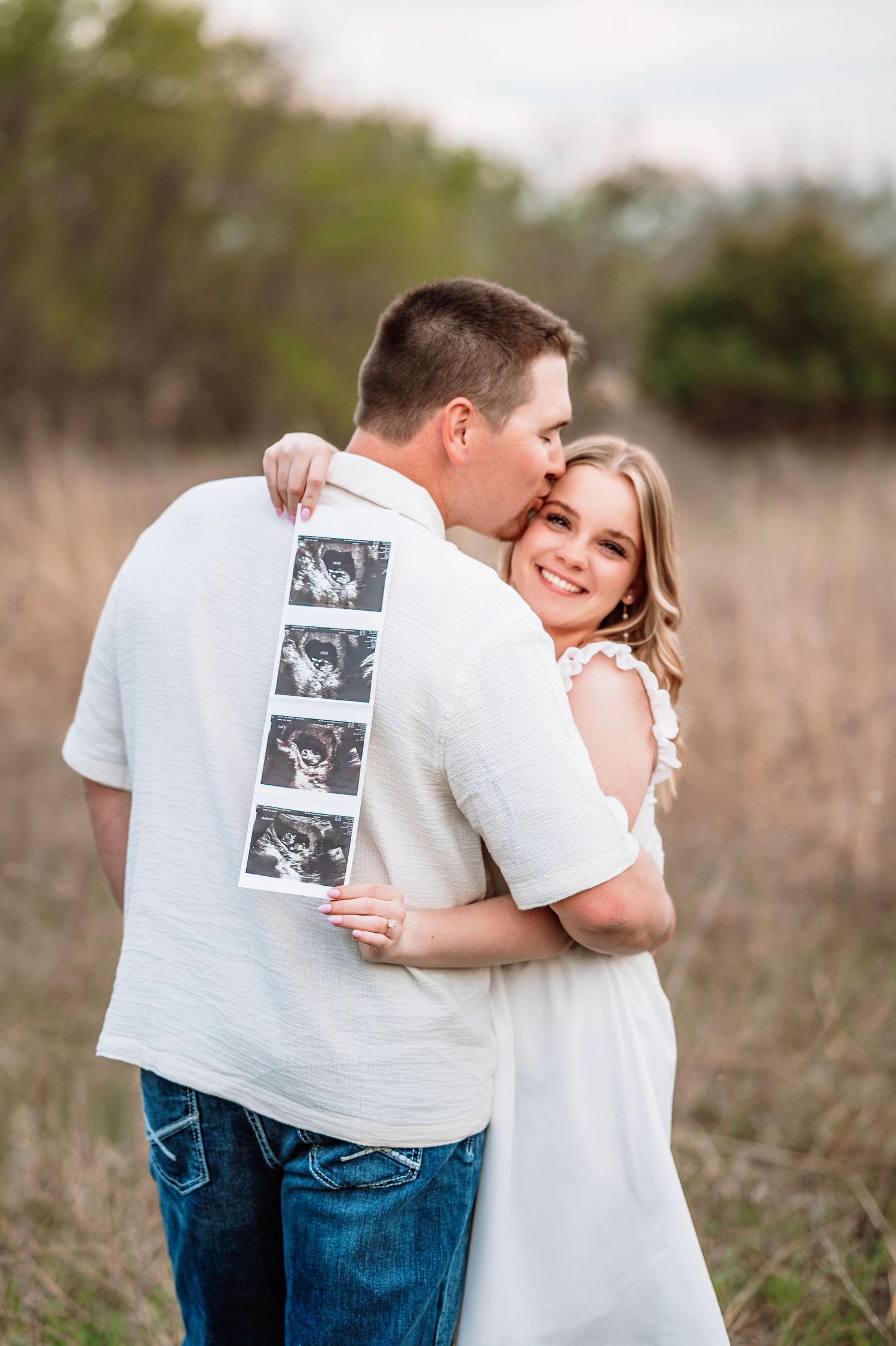 A couple embraces in a field while holding ultrasound photos displayed on the back of a white shirt.