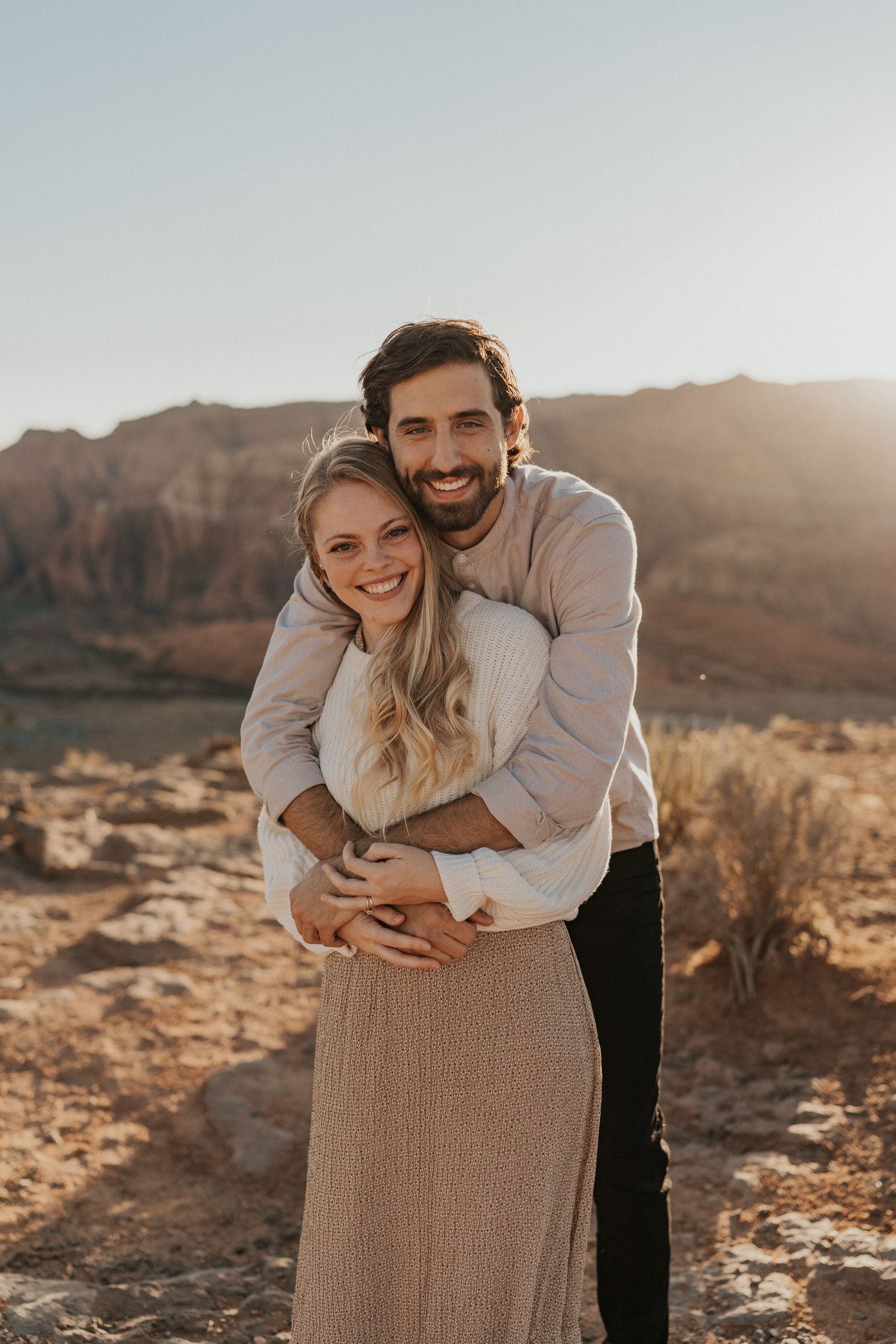 A couple embraces in warm sunlight against a desert mountain backdrop wearing neutral-colored clothing in a romantic pose.