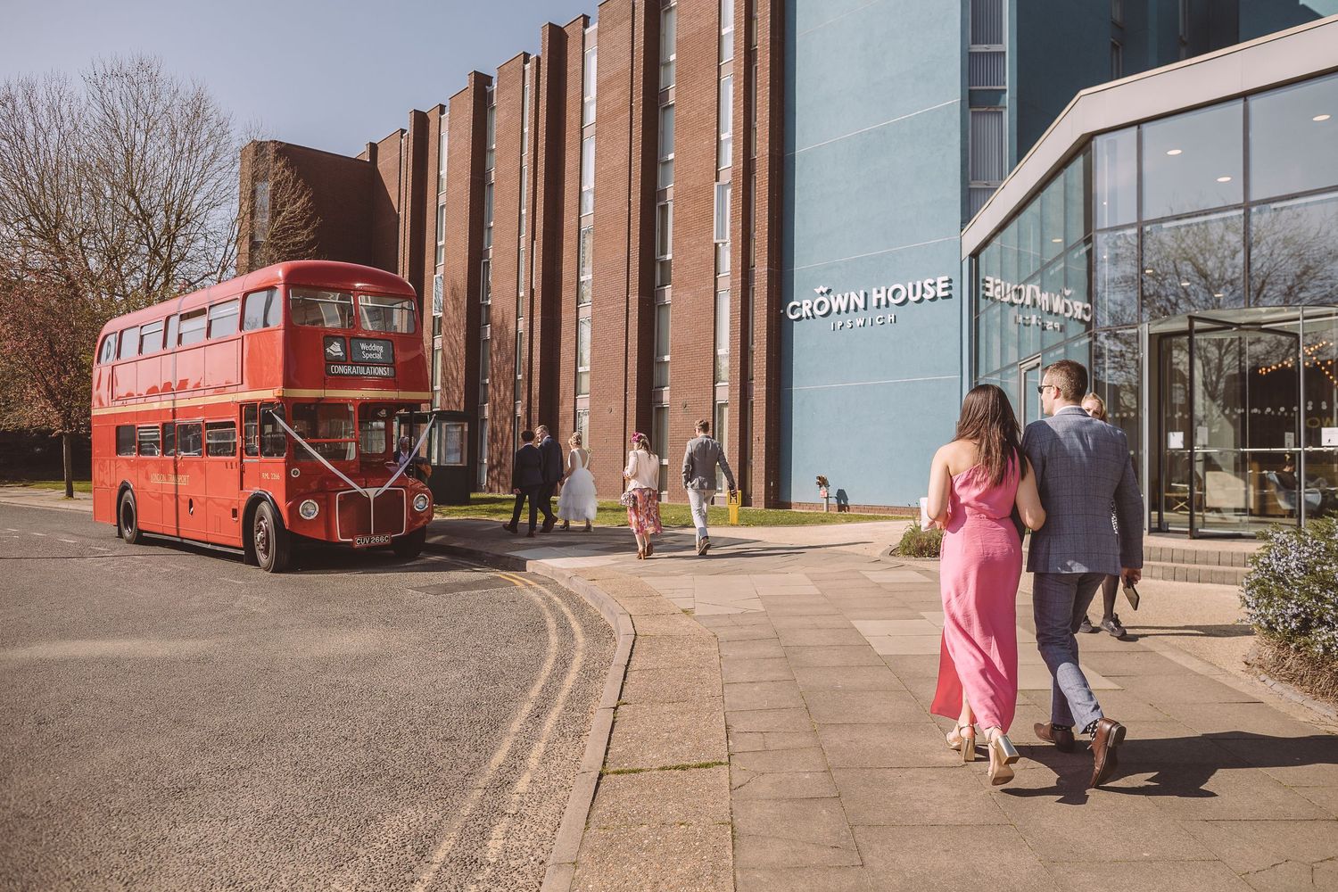 A vintage red double decker bus parked outside Crown House with wedding guests walking towards the bus on a sunny day.